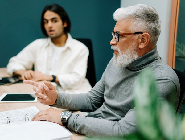 Workplace coaching pre-meeting around a table with a team member in a white shirt and a leader with a grey sweater, grey hair and black glasses. An iPad and folder are on the table.