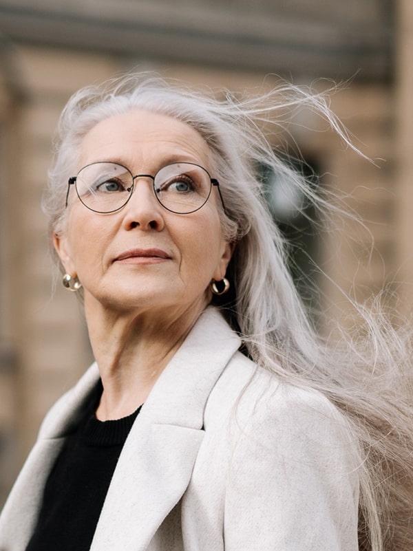 Female leadership coach with long grey hair, glasses and cream blazer looking into the street