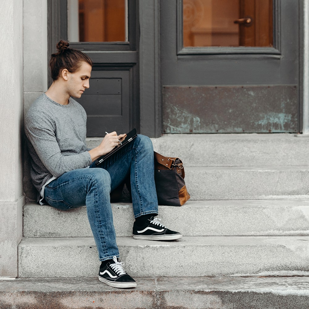 Man in grey t-shirt and jeans sitting on the steps of a building applying for mentor coaching.